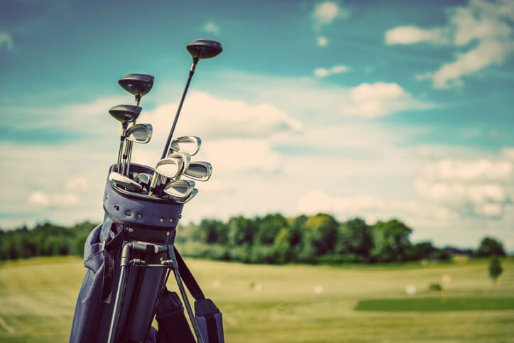 Close up of a golf bag with assorted clubs and a fairway line by trees in the background.
