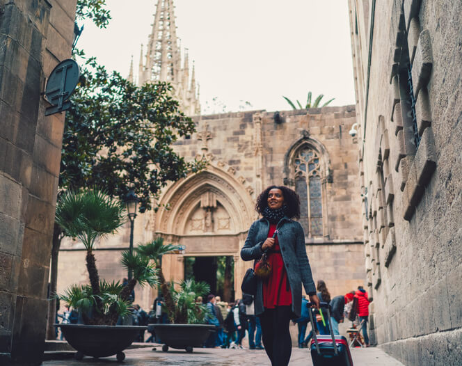 A woman smiling as she pulls a rolling suitcase through the streets of Barcelona, knowing her travel insurance questions have been answered.